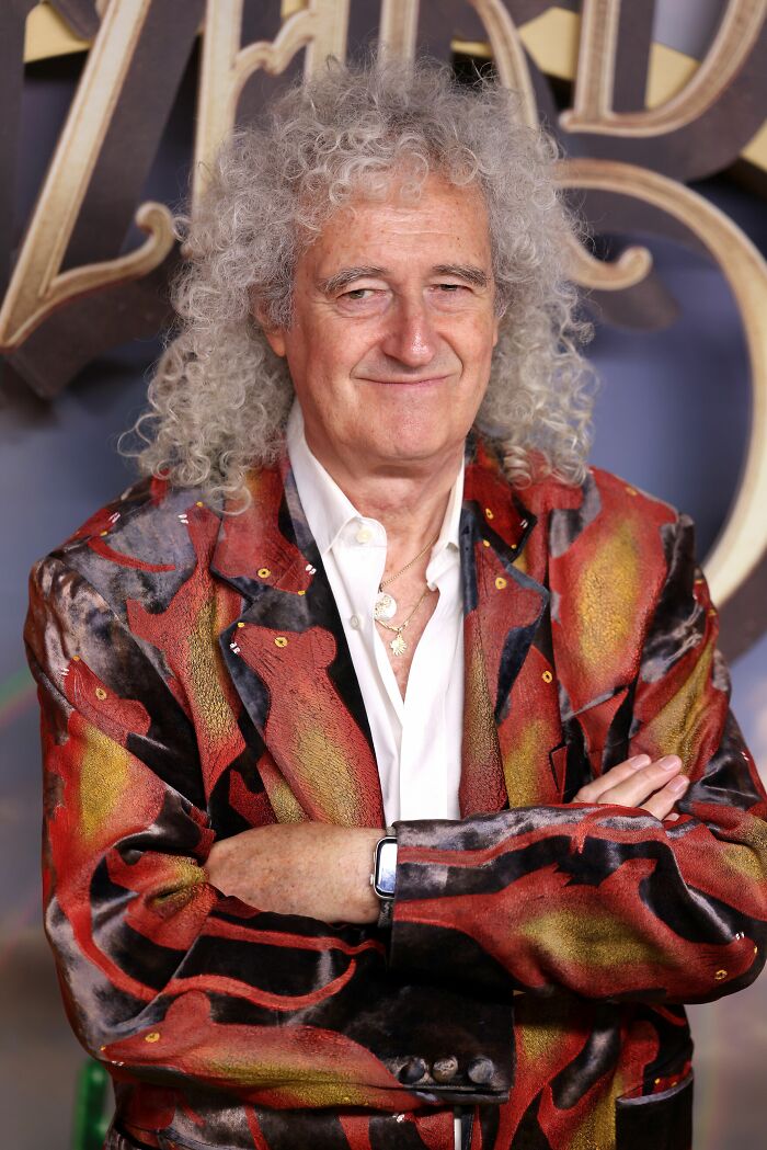 Brian May with curly grey hair, wearing a patterned red, black, and gold jacket over a white shirt, smiling, arms crossed. This image showcases Brian May in an iconic look, highlighting his unique style and personality.