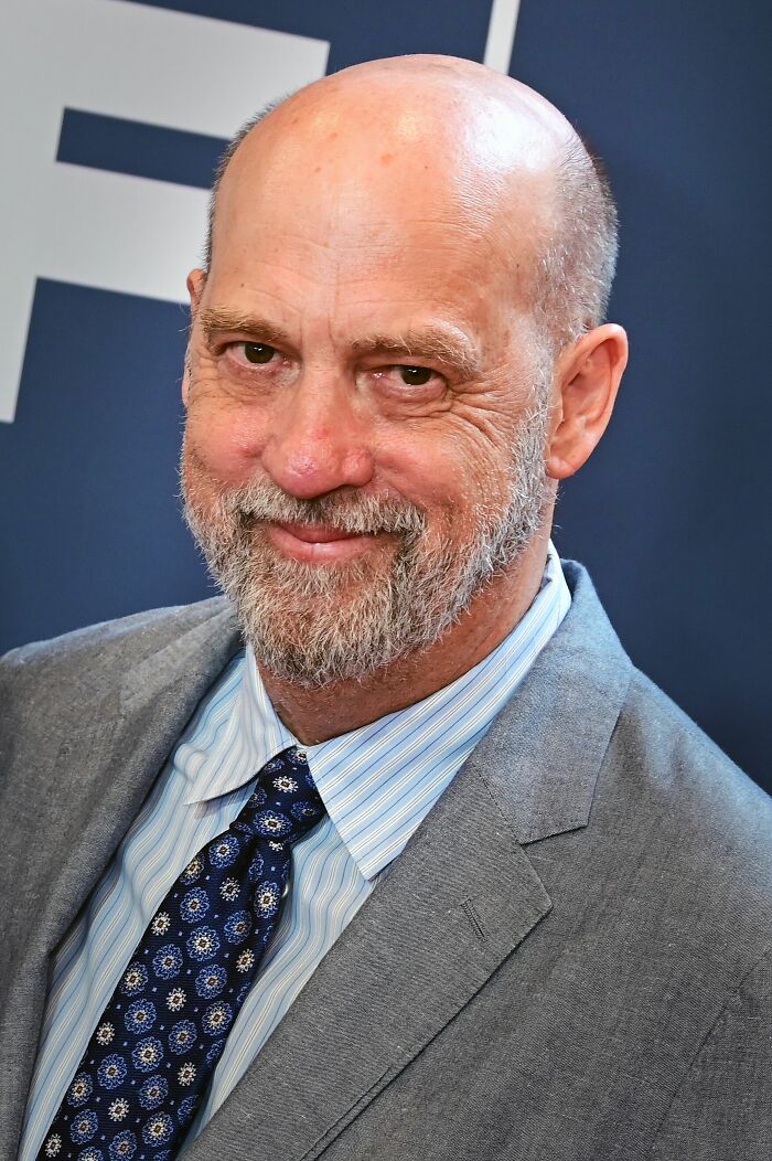 A portrait of Anthony Edwards, a bald man with a gray beard, wearing a gray suit and a patterned blue tie, smiling.
