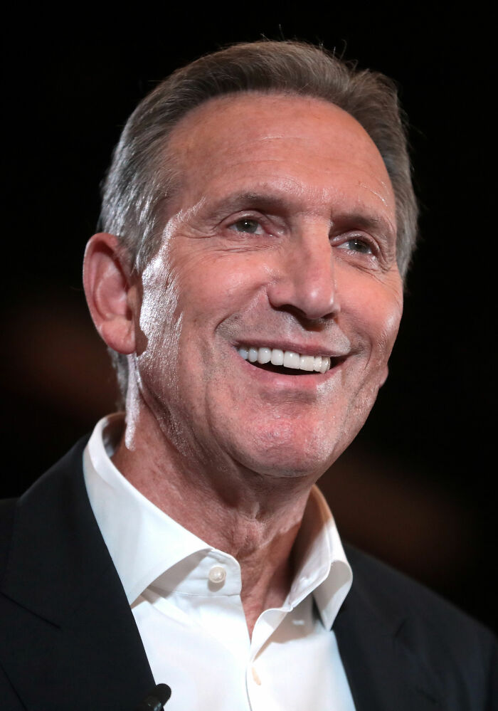 Howard Schultz smiling in a suit. His bio and career highlights include leading Starbucks.