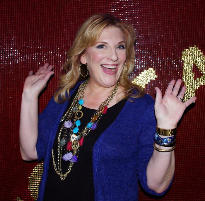 Lisa Lampanelli with blonde hair, a blue cardigan, and colorful necklaces smiles with hands raised. Career Highlights.