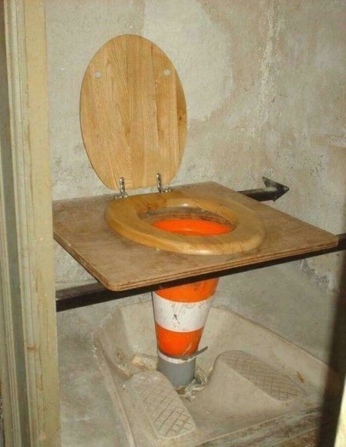 A toilet seat on a wooden plank, supported by an orange traffic cone, a clever solution using random junk.