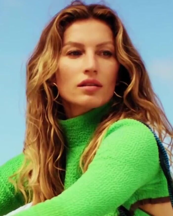 Gisele Bundchen poses for a photo, showcasing her career highlights. She has long, wavy hair and is wearing a green sweater.