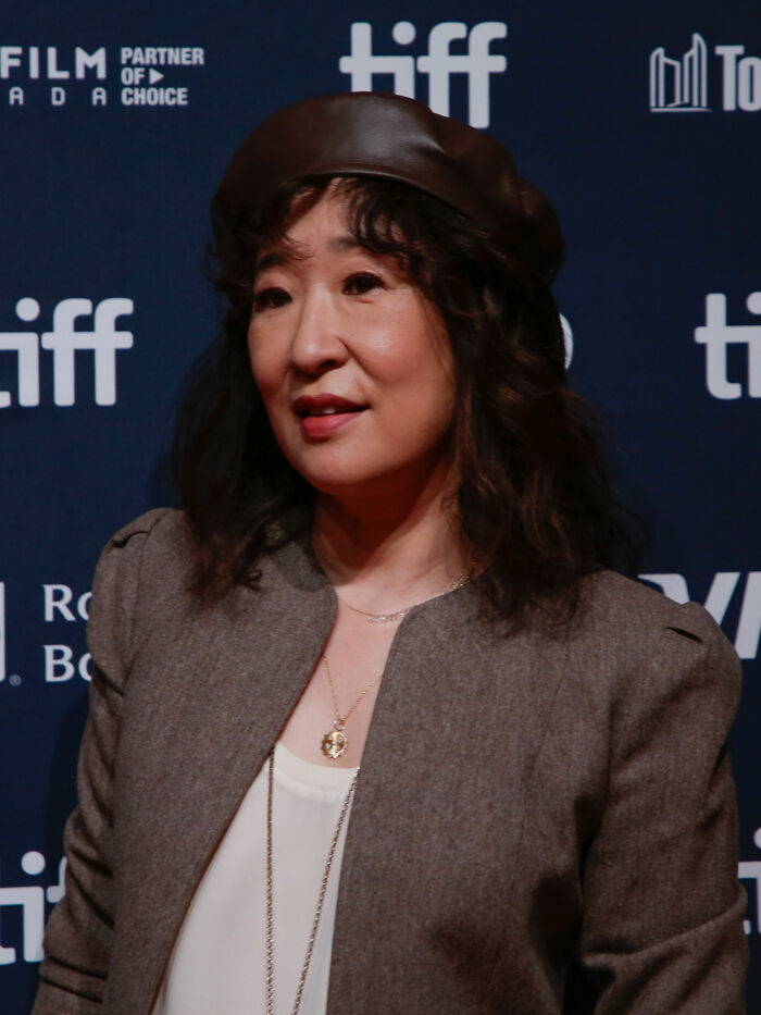 Sandra Oh smiling at an event, wearing a brown beret and a grey blazer, capturing her career highlights.