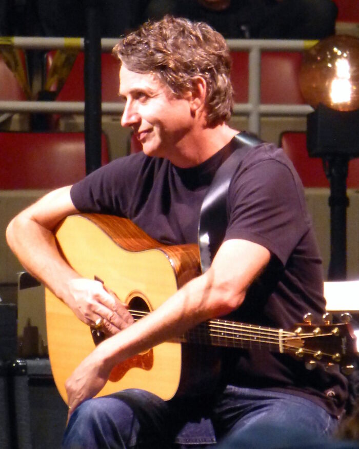 Stone Gossard playing acoustic guitar, showcasing career highlights, with a subtle smile in a live performance setting.