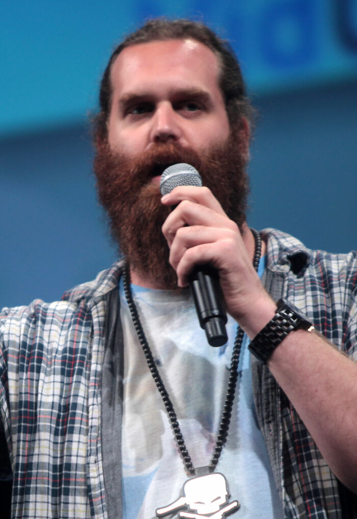 Harley Morenstein with a prominent beard and long hair, speaking into a microphone, highlighting his career.