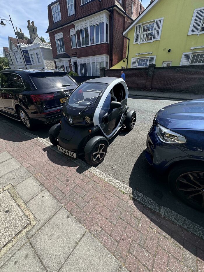 A tiny white and black car is parked partially on the sidewalk next to a larger black SUV, an example of outrageous parking.
