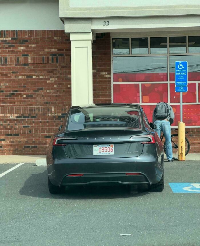 A dark gray Tesla is parked in a handicapped spot, with a person bending over a bicycle, illustrating outrageous parking.