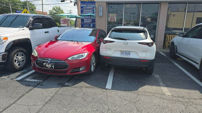 A red Tesla and a white Mazda are parked very close, nearly touching, in a parking lot, an example of outrageous parking.