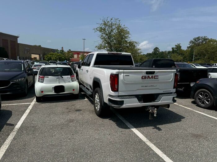 A large white GMC truck is parked across two spaces next to a small white car, displaying outrageous parking.