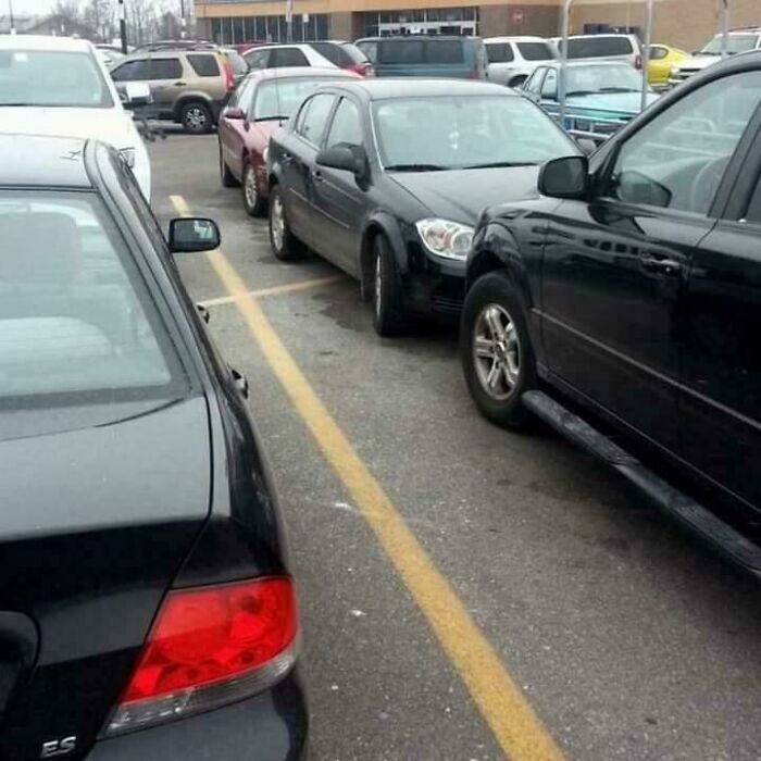 A black car is parked over a yellow line next to other vehicles in a parking lot, highlighting outrageous parking.