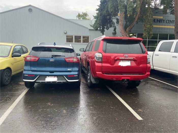 Two cars, a blue Kia and a red Toyota SUV, are parked side-by-side, poorly centered within their outrageous parking spaces.