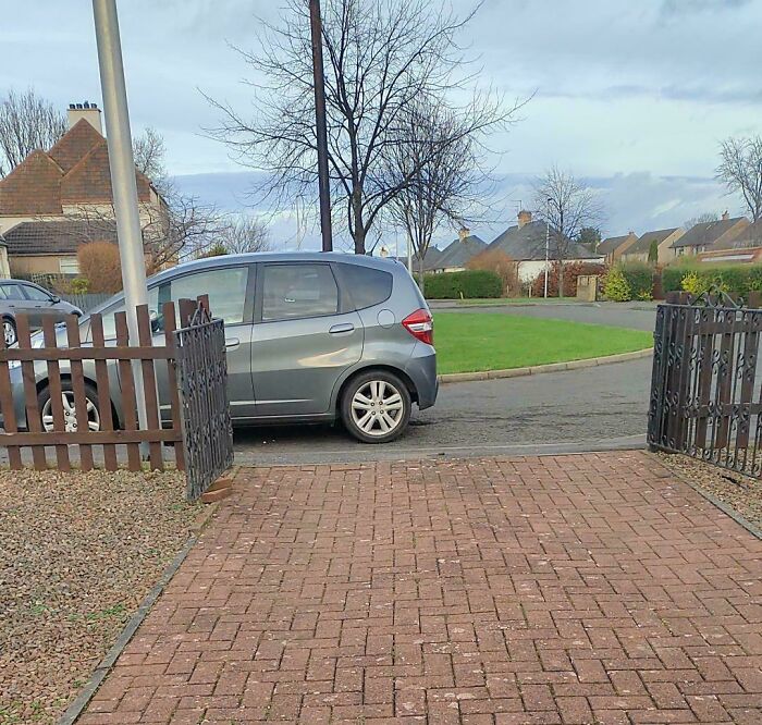 A grey car obstructing a driveway entrance with its poor parking, demonstrating outrageous parking.
