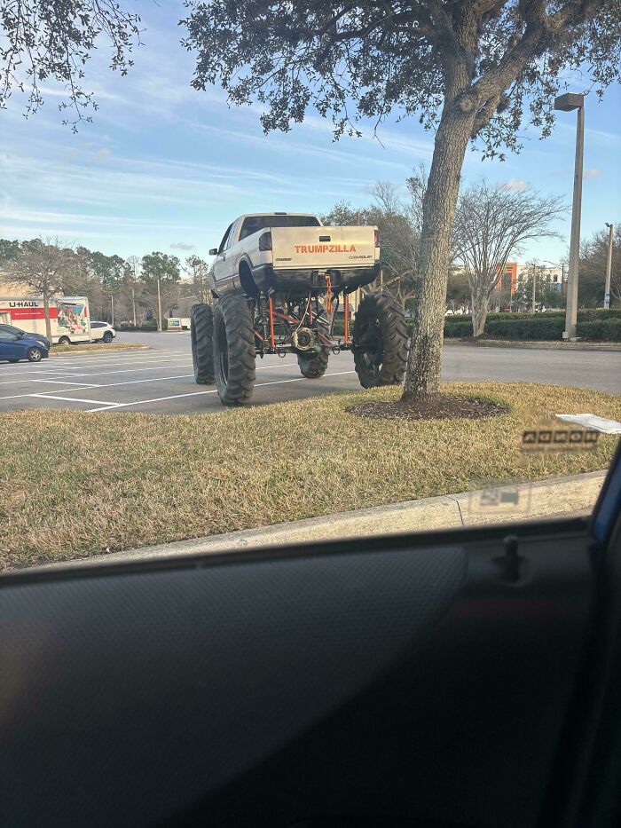 A massive monster truck, labeled TRUMPZILLA, parked outrageously on the grass next to a tree in a parking lot.