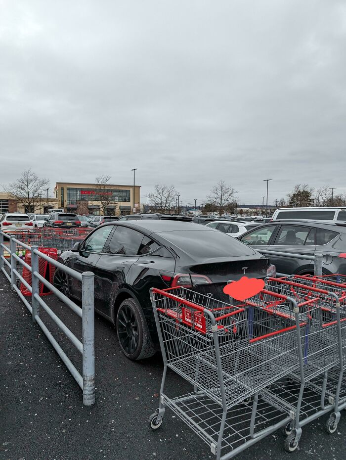 A black Tesla parked outrageously, blocking a shopping cart return area at a store, demonstrating poor parking.