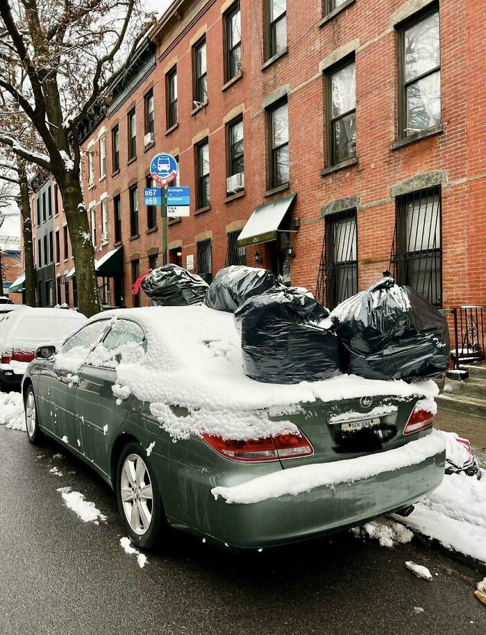 A snow-covered car with several black trash bags on its roof, displaying outrageous parking on a city street.