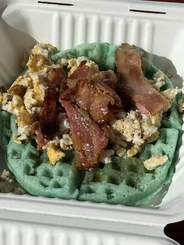 A green waffle topped with scrambled eggs and bacon, a piece of stupid and disgusting food.