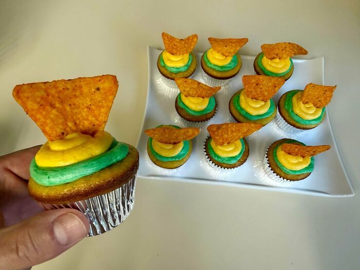 A hand holding a cupcake topped with a Dorito chip, with more such cupcakes, a stupid and disgusting food creation.