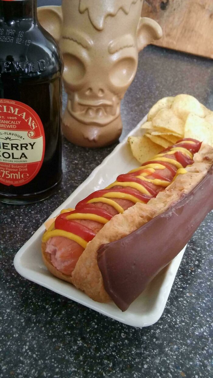 A hot dog with ketchup and mustard next to a chocolate eclair, chips, and a drink, a truly disgusting food combination.