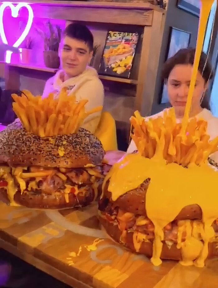 Two customers with stupid food, giant burgers topped with fries and drenched in cheese sauce.