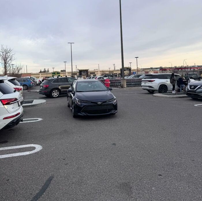 A black car parked diagonally across two spaces in a crowded lot, exemplifying outrageous parking.