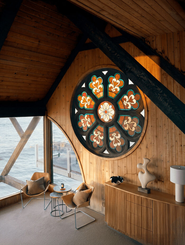 Stunning 1970s and 1980s interior design with a large, colorful stained glass window overlooking the ocean, wood-paneled walls, and two modern chairs.