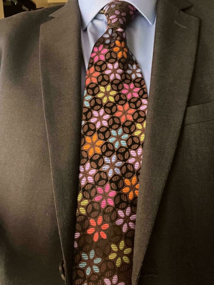 Close-up of a brown tie with a colorful floral pattern, paired with a blue shirt and black suit, reflecting 1970s and 1980s interior design.
