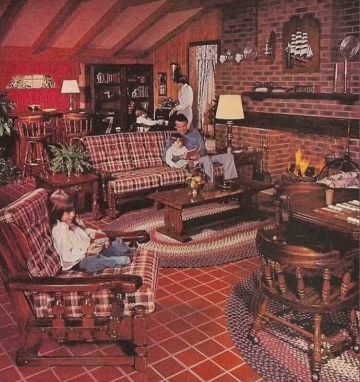 A family relaxing in a cozy living room with a brick fireplace, plaid furniture, and red tile flooring, exemplifying 1970s and 1980s interior design.