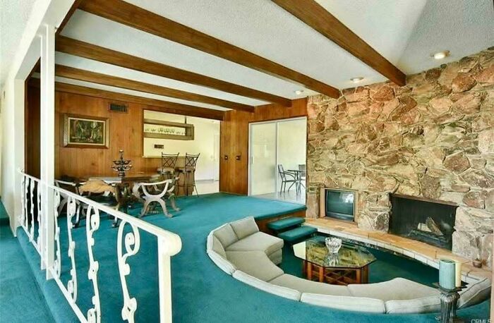 Sunken living room with a turquoise carpet, stone fireplace, wooden beams, and a view of a dining area, epitomizing 1970s and 1980s interior design.