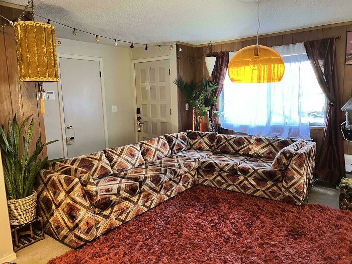 A cozy living room featuring a large patterned sectional sofa, characteristic of 1970s and 1980s interior design.