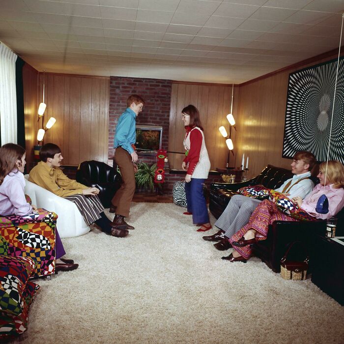 A group of people relaxing in a living room, showcasing 1970s and 1980s interior design with bold patterns and wood paneling.