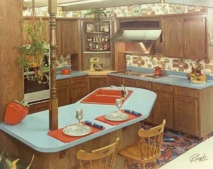 A 1980s interior design kitchen featuring blue countertops, wood cabinetry, and a patterned wallpaper.