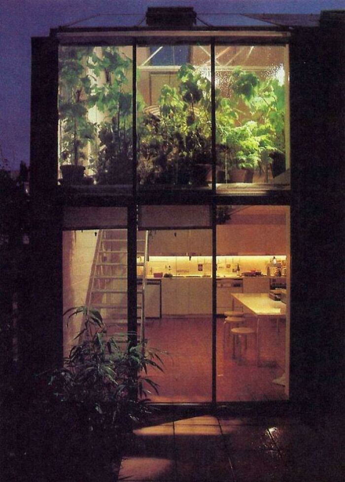 An exterior view of a modern house highlighting 1970s interior design with large windows, lush plants, and a visible kitchen.