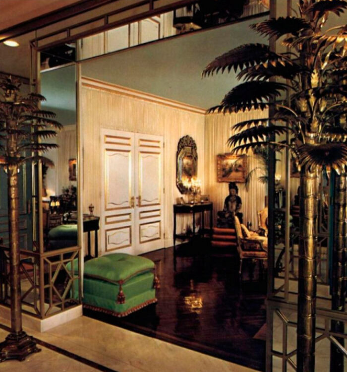 An elegant room with mirrored walls, golden palm trees, and a green ottoman showcases 1970s and 1980s interior design.