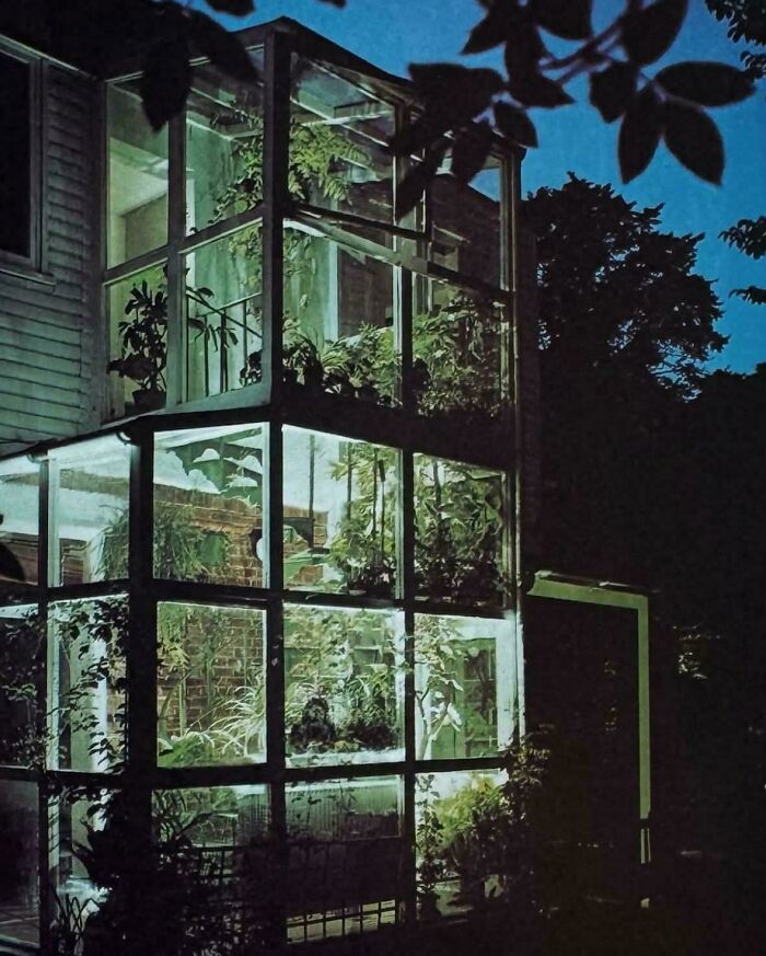 A multi-story glass structure filled with plants, illuminated at night, reflecting 1970s and 1980s interior design.