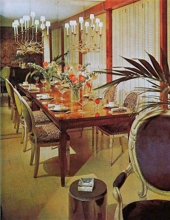 A formal 1970s and 1980s interior design dining room with a long wooden table, patterned chairs, and two chandeliers.