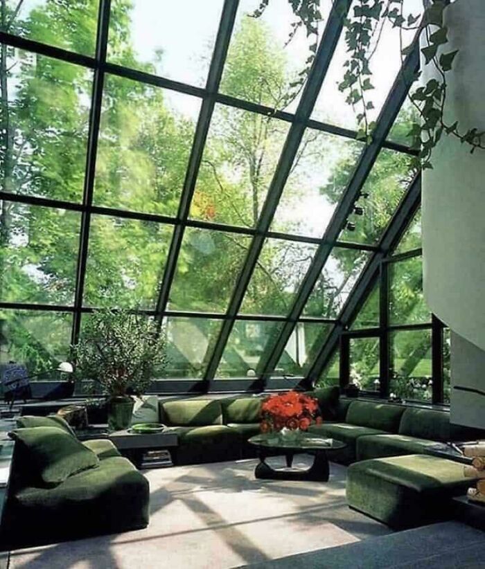 A living room with an expansive glass wall offering a view of lush trees, exemplifying 1970s and 1980s interior design with green sofas.