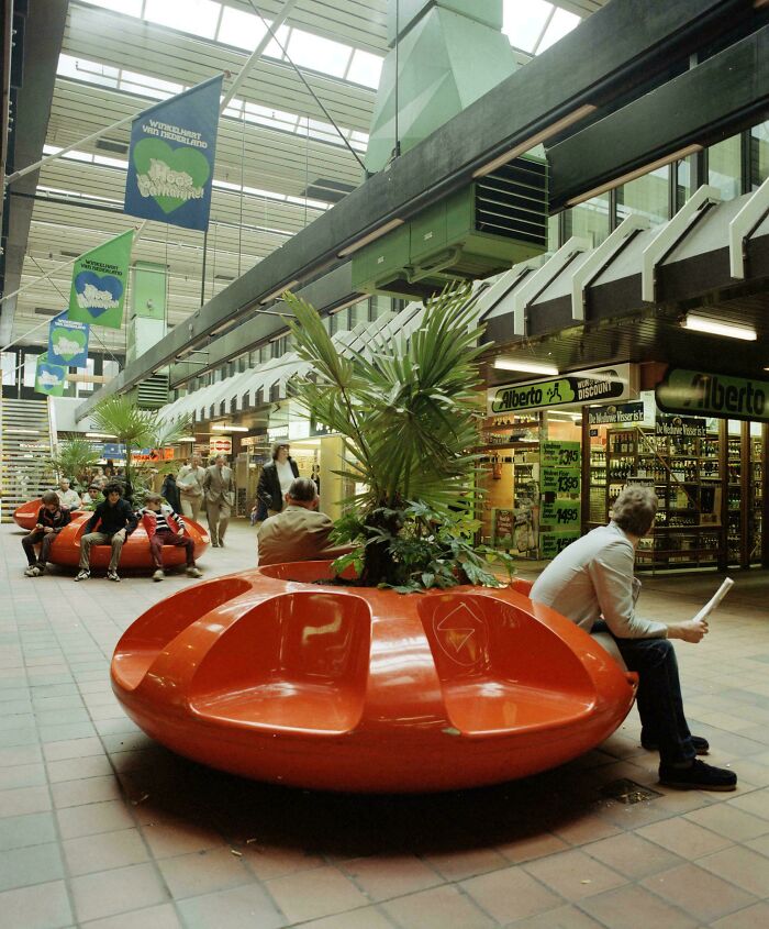 People sitting on orange benches inside a shopping mall, an example of 1970s and 1980s interior design.