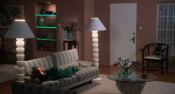 A person relaxing on a striped couch in a living room, highlighting 1970s and 1980s interior design with unique lamps and neon accents.