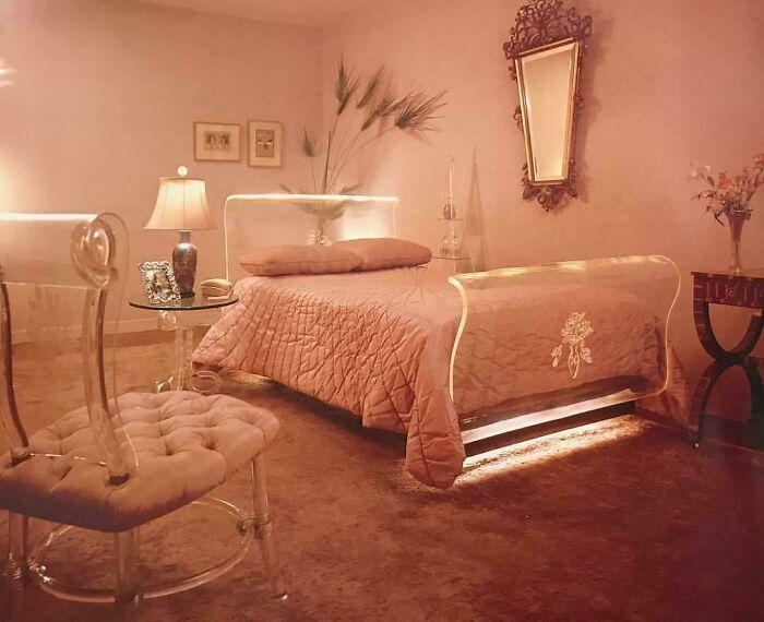 A stylish 1970s bedroom interior design with a pink bed, mirrored headboard, and clear acrylic furniture.