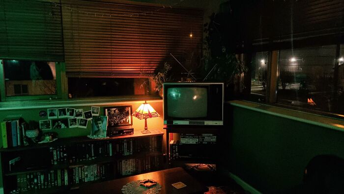 A dim room showcasing 1970s and 1980s interior design with a vintage TV, wooden blinds, and a bookshelf.