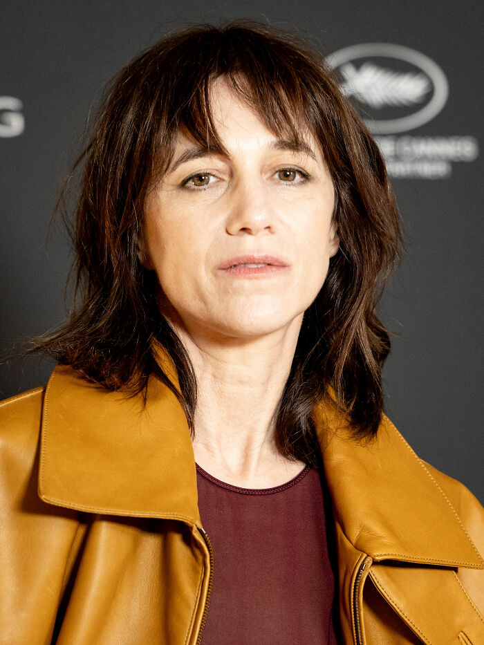 A close-up shot of Charlotte Gainsbourg, an actress with dark hair and a light brown jacket, looking directly at the camera.