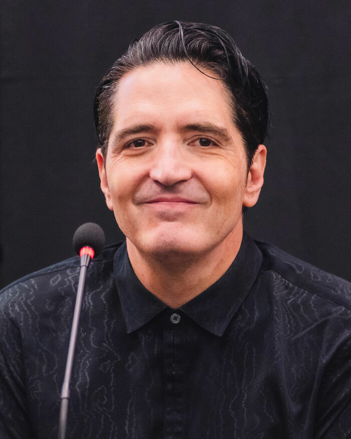 Smiling David Dastmalchian looking directly at the camera with a microphone in the foreground, highlighting his career.