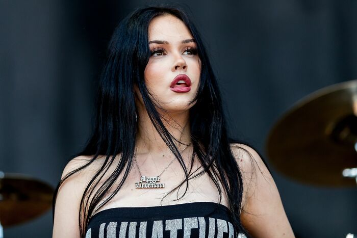 Maggie Lindemann, with long dark hair, wearing a black top and a nameplate necklace, looks upward during a performance.
