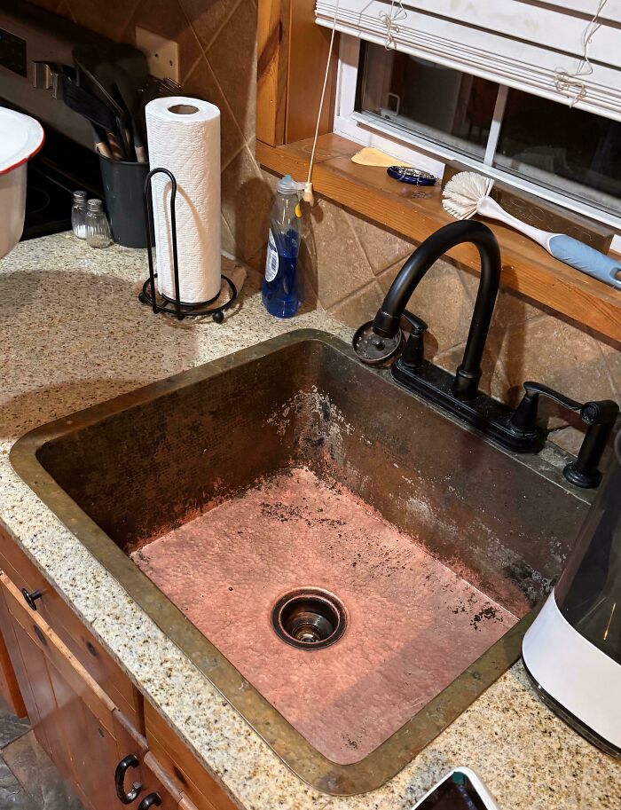 A dirty kitchen sink that needs fixing things. The sink is made of copper and has a dark patina.