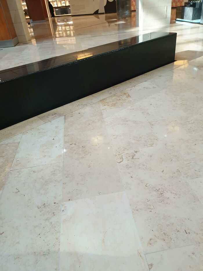 A long, black bench in a public area with a highly reflective floor, highlighting a construction fail.