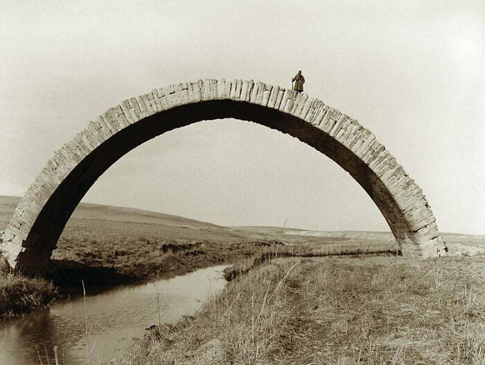 Historical photos: A lone figure stands atop a stone arch bridge spanning a narrow river in a vast, open landscape.