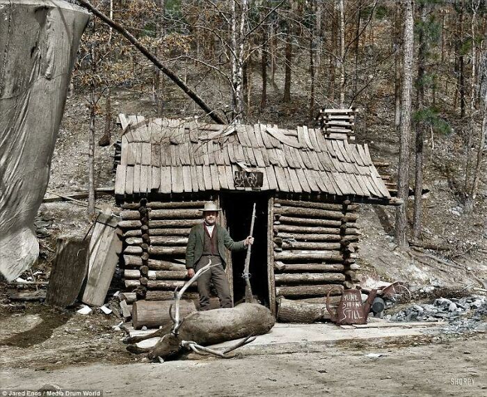 Historical photos: A man standing proudly with his hunting rifle and a k****d deer outside a rustic log cabin in the woods.