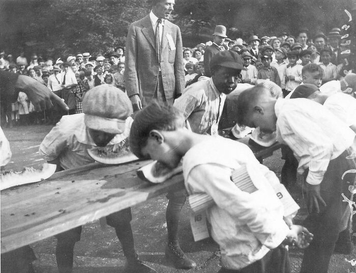 Historical photos: Boys participating in a watermelon eating contest with an adult watching on a summer day.