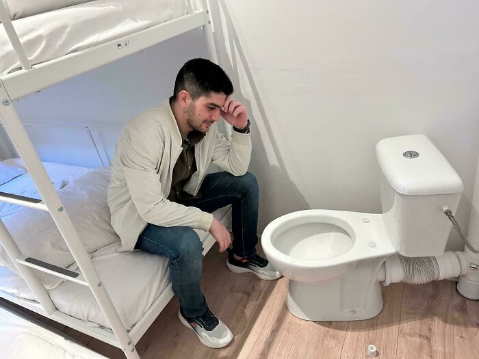 A man looking distressed next to a toilet awkwardly placed beside bunk beds, a notable construction fail.
