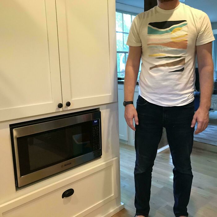 A man standing next to a microwave installed too low in a kitchen cabinet, highlighting construction fails in kitchen design.
